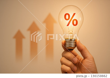 Hand holding glowing light bulb with red percentage symbol inside and rising upward arrows in blurred background, symbolizing investment growth and interest rate increase 130158662