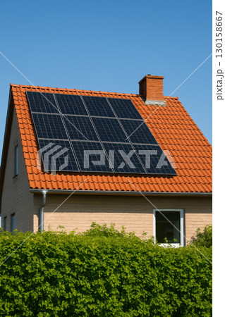 House with red tiled roof covered in black solar panels and lush green hedge in foreground under clear blue sky on sunny day 130158667