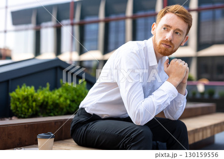Stylish redhead male in white shirt sitting on bench in city district enjoying warm weather 130159556