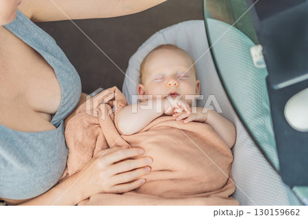 Multitasking mom works on a laptop at home while her newborn baby sleeps peacefully in a cozy baby nest on her lap. Concept of remote work, motherhood, and balancing family and career 130159662
