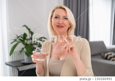 Blonde woman holding pill in one hand and glass of water in the other, preparing to take supplement Blonde woman holding pill in one hand and glass of water in the other, preparing to take supplement 130160491