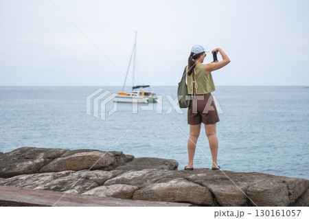 Asian woman taking photo of sailboat on ocean from rocky shore during daytime Asian woman taking photo of sailboat on ocean from rocky shore during daytime 130161057