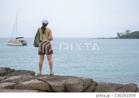 Asian woman standing on rocky shore looking at sailboat on calm sea Asian woman standing on rocky shore looking at sailboat on calm sea 130161059