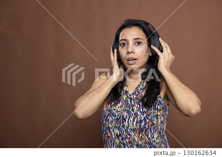 Young female adult wearing casual clothing and wireless headphones, dancing to rhythm in modern studio. Pretty indian woman looking at camera and enjoying her favorite tunes against brown background. Young female adult wearing casual clothing and wireless headphones, dancing to rhythm in modern studio. Pretty indian woman looking at camera and enjoying her favorite tunes against brown background. 130162634