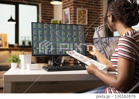 Focused female entrepreneur takes notes on clipboard during active trading session in brick wall room. African American woman works from home, studying stock market data on desktop monitor. 130162679