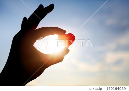A silhouette of a woman holding a red heart, a symbol of precious life and love, against a blue sky and bright sunlight background. 130164089