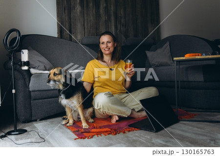 Healthy lifestyle at home. Smiling woman with juice and dog, sitting on the floor in cozy living room. Concept of biohacking, vegetarian habits, pet care, and emotional balance 130165763