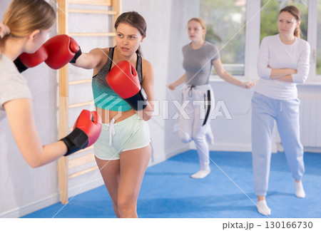 Two teenage girls in sportswear are engaged in boxing sparring in the gym under guidance of trainer in gym 130166730