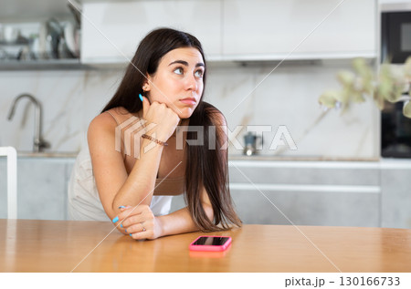 Young woman leaning on kitchen table waiting for call on smartphone Young woman leaning on kitchen table waiting for call on smartphone 130166733