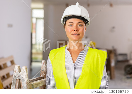 Confident female builder in vest and hard hat standing at construction site 130166739