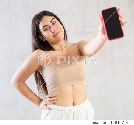 Young woman showing her mobile phone standing against wall 130167160