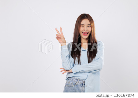 Victory fingers raised in joyful expression, celebrating a moment of victory with a smiling Asian woman. Peaceful gesture, two fingers up in a cheerful V sign isolated on white background. 130167874