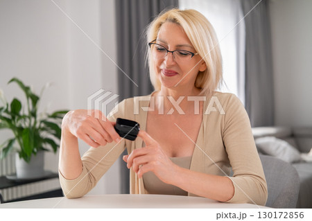 Smiling blonde woman in living room measuring heart rate with oximeter. 130172856