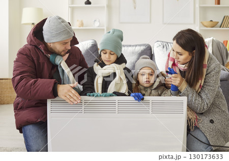 Family in warm clothes sitting by electric heater, trying to stay warm in cold room during winter. 130173633