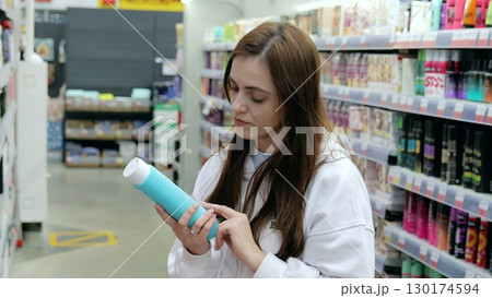 Young woman shopping for cosmetics, attentively checking ingredients and information on shampoo bottle in a brightly lit supermarket aisle 130174594