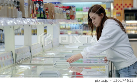 Young woman opening freezer in supermarket, choosing frozen products while shopping for groceries in refrigerated section Young woman opening freezer in supermarket, choosing frozen products while shopping for groceries in refrigerated section 130174613