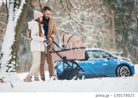 With pram. Happy couple having a walk in winter forest. Blue car is parked 130178025