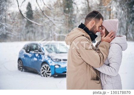 Standing and embracing each other. Happy couple having a walk in winter forest. Blue car is parked 130178070