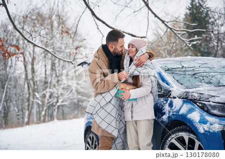 With warm towel. Happy couple having a walk in winter forest. Blue car is parked 130178080
