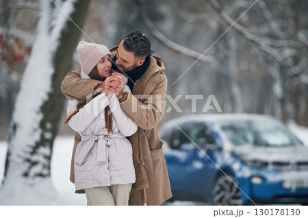 Standing and embracing. Happy couple having a walk in winter forest. Blue car is parked 130178130