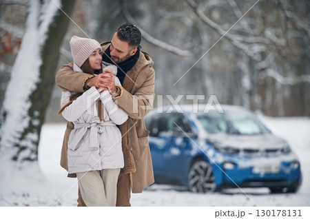 Standing and embracing. Happy couple having a walk in winter forest. Blue car is parked 130178131