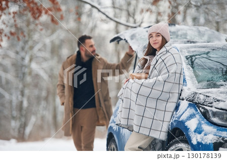 Man is holding the towel to warm the woman. Happy couple having a walk in winter forest. Blue car is parked 130178139