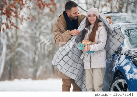 Man is holding the towel to warm the woman. Happy couple having a walk in winter forest. Blue car is parked 130178141