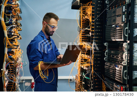 With laptop in hands. Young man is working with internet equipment and wires in server room 130178430