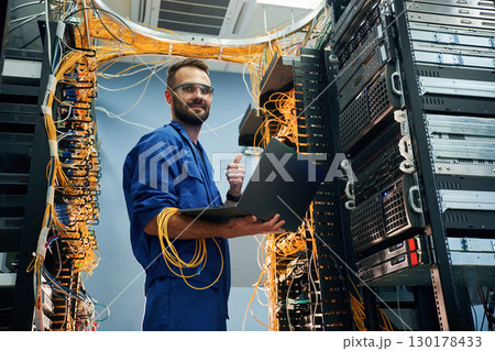 Silver colored laptop in hands. Young man is working with internet equipment and wires in server room Silver colored laptop in hands. Young man is working with internet equipment and wires in server room 130178433