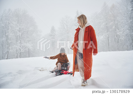 A woman pulls a sled. Mother and her daughter is on the winter meadow and forest A woman pulls a sled. Mother and her daughter is on the winter meadow and forest 130178929