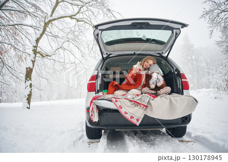 Sitting, with warm drink. Woman with her daughter is on the trunk of a car in the winter meadow and forest 130178945