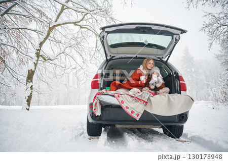 Sitting, with warm drink. Woman with her daughter is on the trunk of a car in the winter meadow and forest Sitting, with warm drink. Woman with her daughter is on the trunk of a car in the winter meadow and forest 130178948