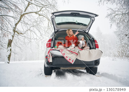 On the towel, with warm drink. Woman with her daughter is on the trunk of a car in the winter meadow and forest On the towel, with warm drink. Woman with her daughter is on the trunk of a car in the winter meadow and forest 130178949