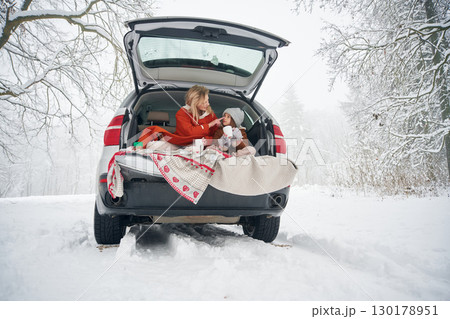 On the towel, with warm drink. Woman with her daughter is on the trunk of a car in the winter meadow and forest 130178951