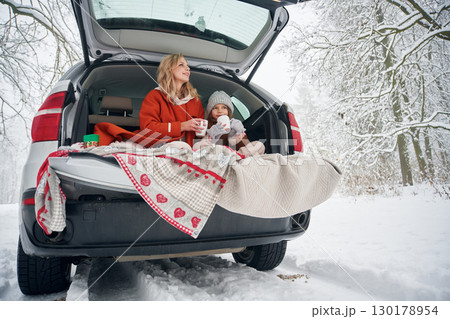 Drinking from the cup. Woman with her daughter is on the trunk of a car in the winter meadow and forest 130178954
