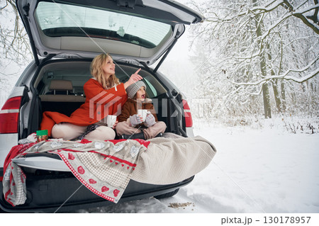 Drinking from the cup. Woman with her daughter is on the trunk of a car in the winter meadow and forest Drinking from the cup. Woman with her daughter is on the trunk of a car in the winter meadow and forest 130178957