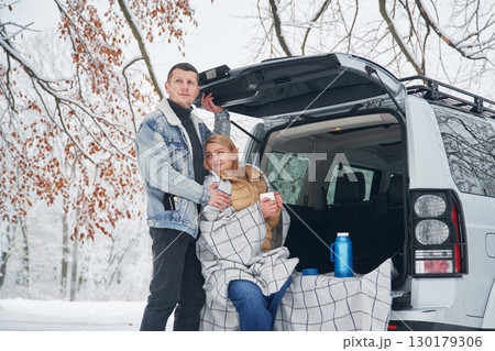 Woman is sitting in the trunk. Beautiful couple standing near the car in the winter forest 130179306