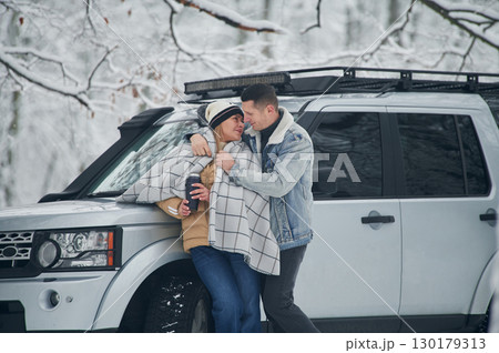 Man is covering woman with warm blanket. Beautiful couple standing near the car in the winter forest Man is covering woman with warm blanket. Beautiful couple standing near the car in the winter forest 130179313