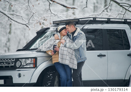 Man is covering woman with warm blanket. Beautiful couple standing near the car in the winter forest Man is covering woman with warm blanket. Beautiful couple standing near the car in the winter forest 130179314