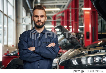 With arms crossed, standing. Car repairman is in the garage with automobile With arms crossed, standing. Car repairman is in the garage with automobile 130179467