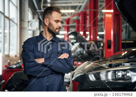 With arms crossed, standing. Car repairman is in the garage with automobile With arms crossed, standing. Car repairman is in the garage with automobile 130179468