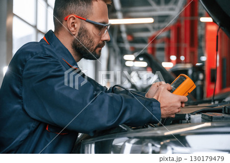 Yellow colored device for measuring electricity is in hands. Car repairman is in the garage with automobile 130179479