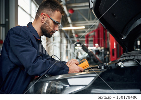 Yellow colored device for measuring electricity is in hands. Car repairman is in the garage with automobile Yellow colored device for measuring electricity is in hands. Car repairman is in the garage with automobile 130179480