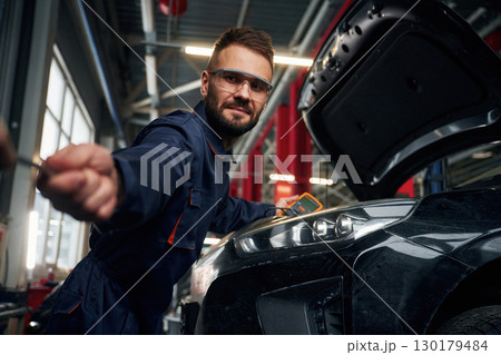 Looking at the camera. Car repairman is in the garage with automobile 130179484