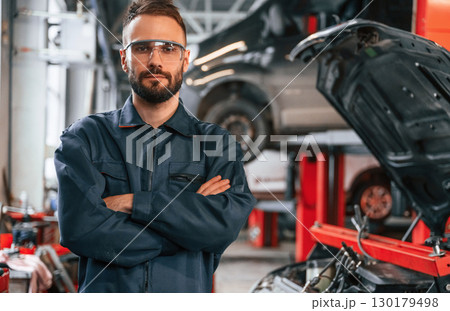 With arms crossed, standing. Car repairman is in the garage with automobile 130179498