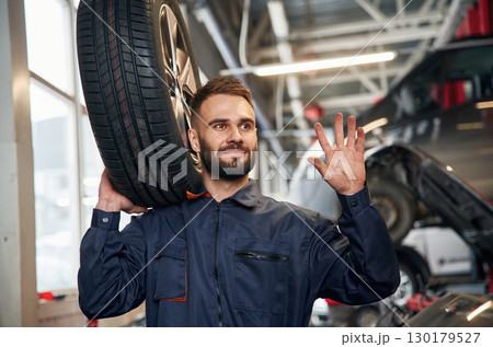 Holding a tire. Car repairman is in the garage with automobile 130179527