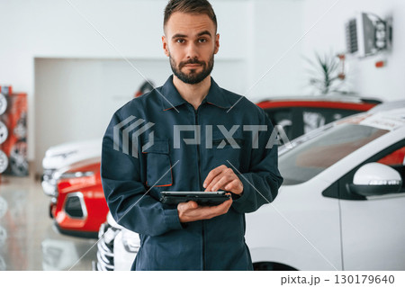 Standing and holding tablet in hands. Car repairman is in the garage with automobile Standing and holding tablet in hands. Car repairman is in the garage with automobile 130179640