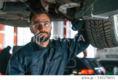 In protective eyewear, under vehicle. Car repairman is in the garage with automobile In protective eyewear, under vehicle. Car repairman is in the garage with automobile 130179651