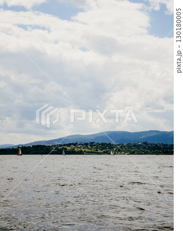 Sailboats are peacefully navigating lake on partly cloudy day, with mountains and small village visible in background, creating serene and picturesque scene. Sailboats gliding across calm lake waters Sailboats are peacefully navigating lake on partly cloudy day, with mountains and small village visible in background, creating serene and picturesque scene. Sailboats gliding across calm lake waters 130180005