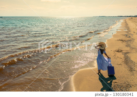 Young happy woman in a long pink dress stands on the beach with a travel suitcase in summer. The woman holds a straw hat. Summertime, travel, and happy holiday concept Young happy woman in a long pink dress stands on the beach with a travel suitcase in summer. The woman holds a straw hat. Summertime, travel, and happy holiday concept 130180341